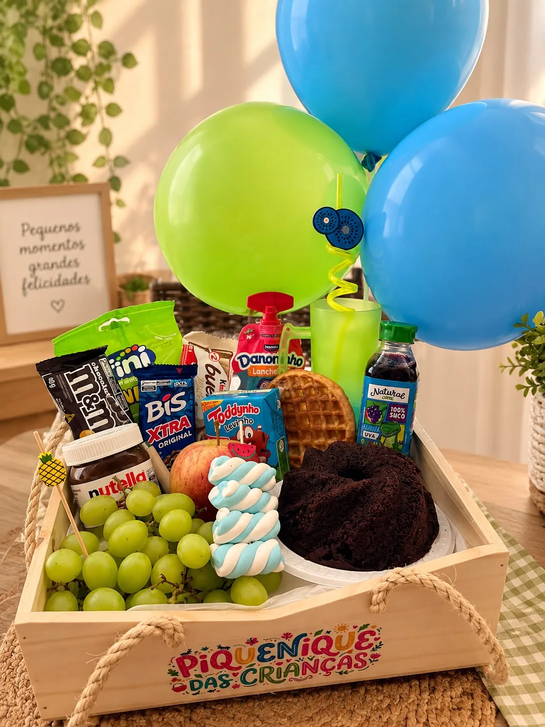 esta de piquenique infantil em caixa de madeira com alças de corda, decorada com balões coloridos em tons de verde e azul, contendo potinho de Nutella, chocolates M&Ms e Bis, wafles, mini bolo de choc